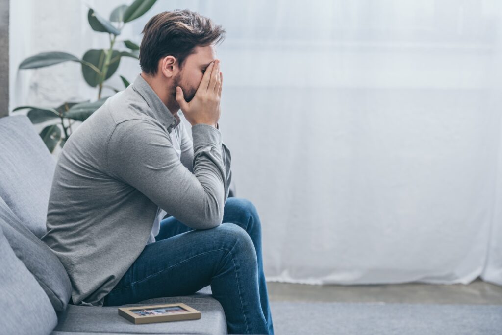 man sitting on grey couch with photo in frame and crying at home, grieving disorder concept